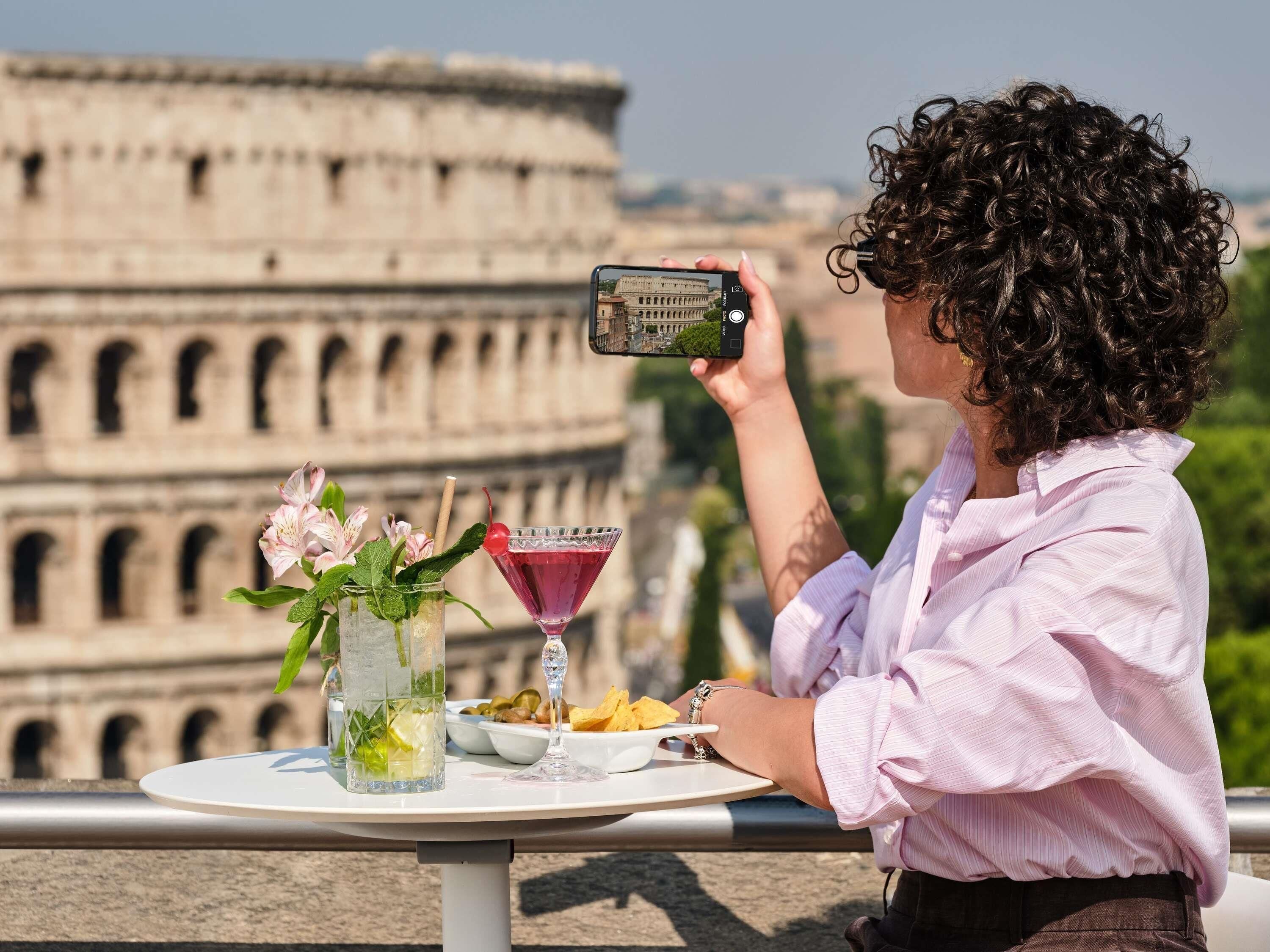 A person taking a photo of the Colosseum with a table in front of them, containing a drink, flowers, and snacks.