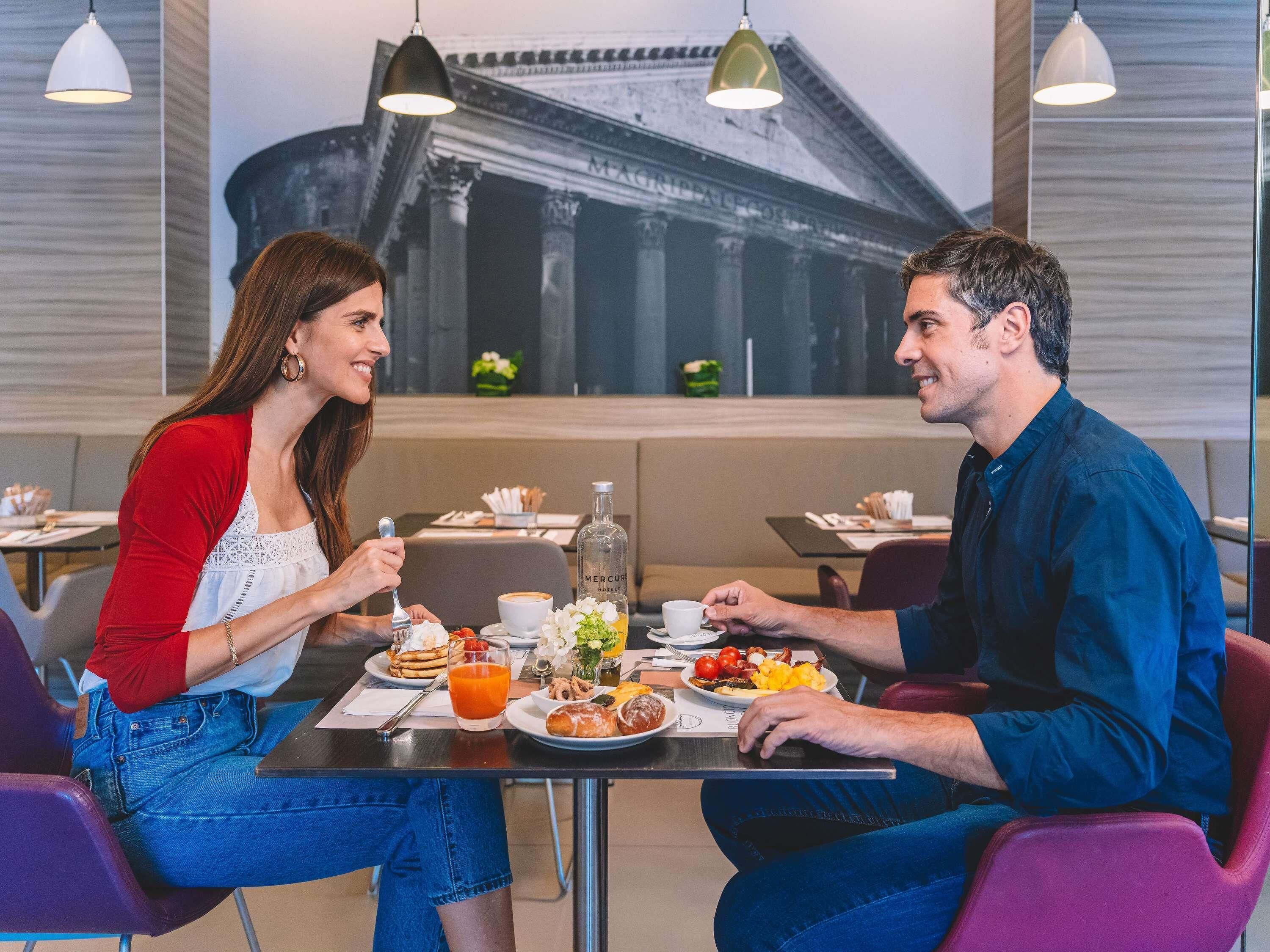 Dos personas cenando en una mesa con un mural de un paisaje urbano al fondo.