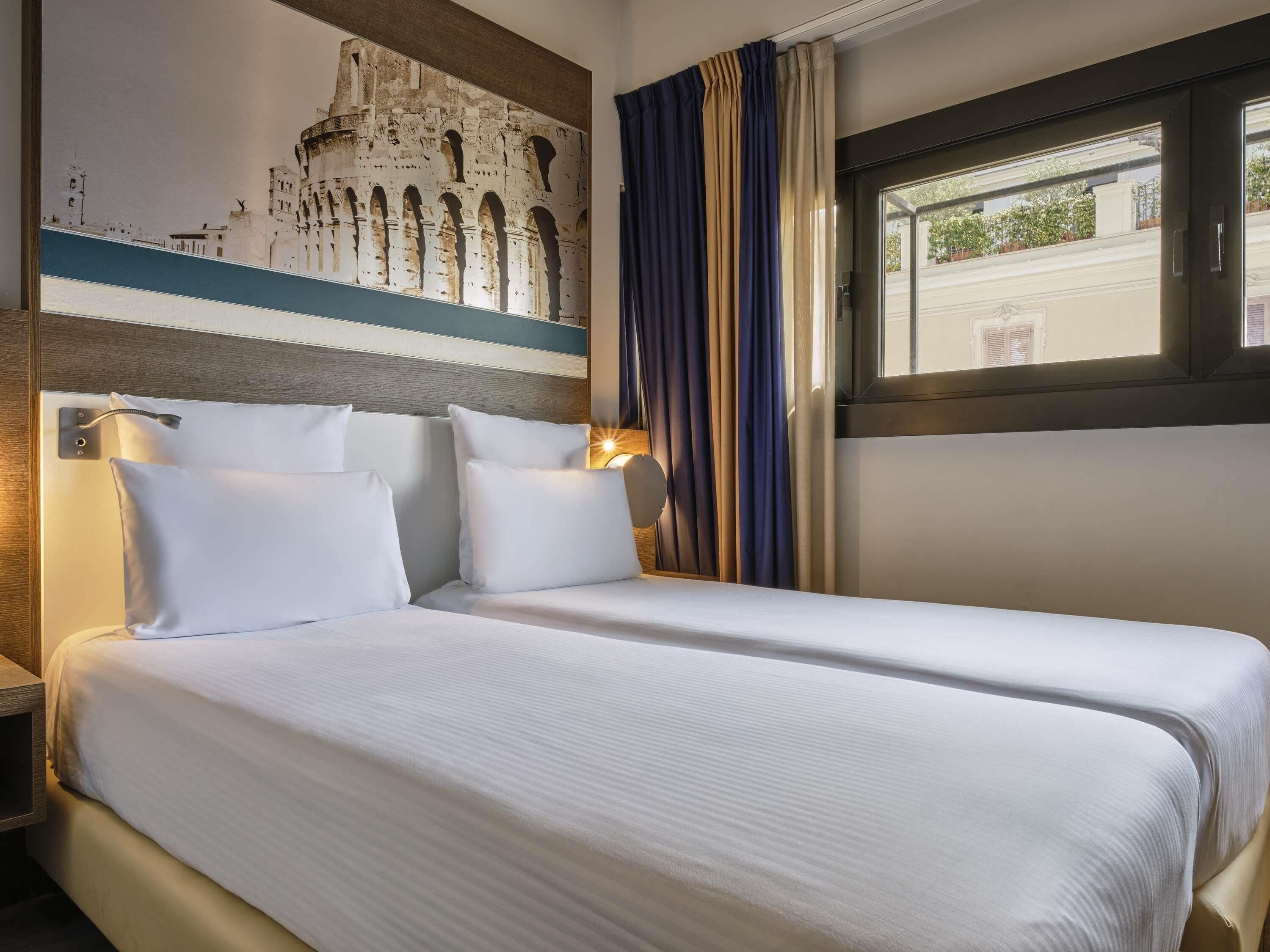 Quarto de hotel com duas camas, um mural grande na parede, uma janela com cortinas e uma mesa de cabeceira com abajur.