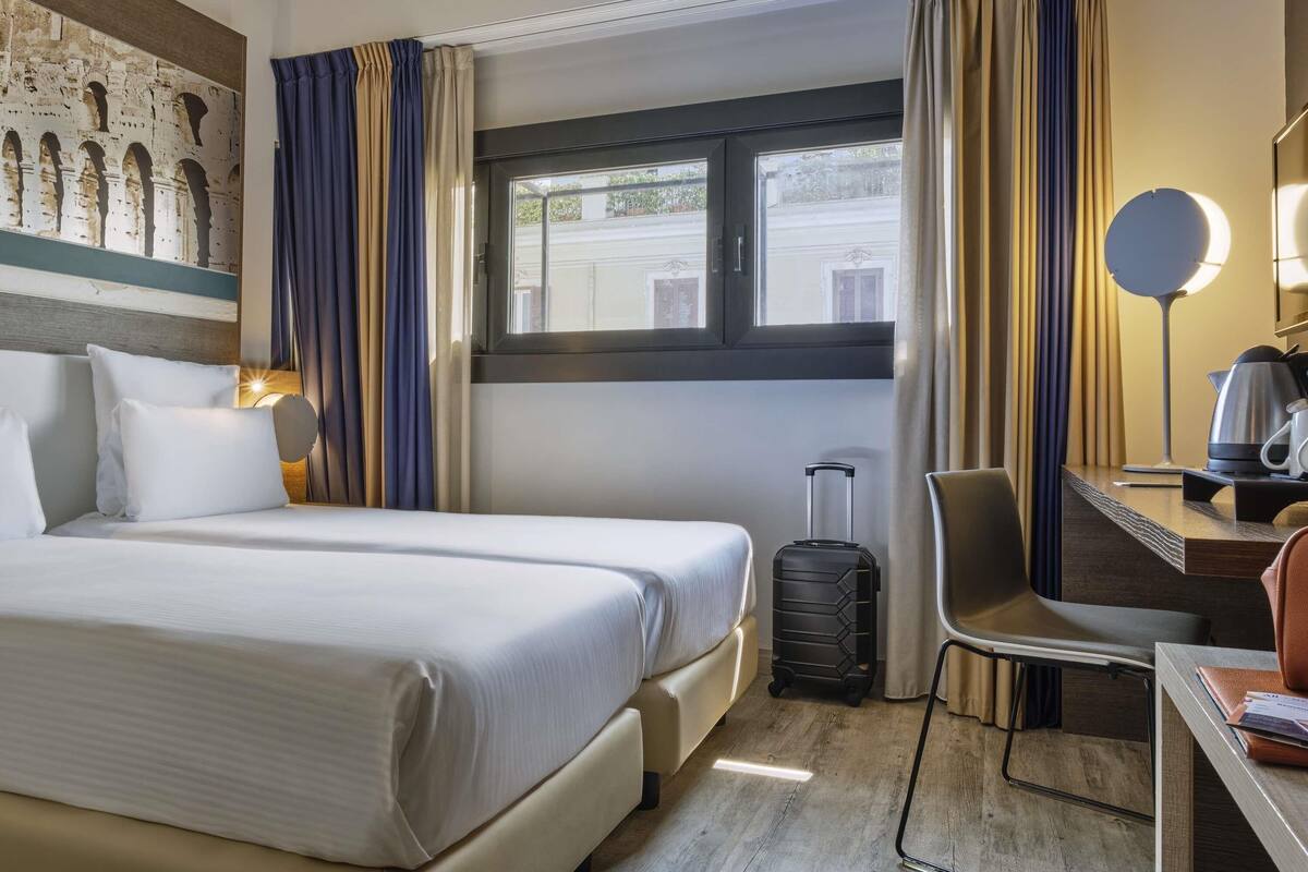 Une chambre d’hôtel avec deux lits, un bureau avec une lampe, une chaise, une valise et une vue sur la rue par une fenêtre.