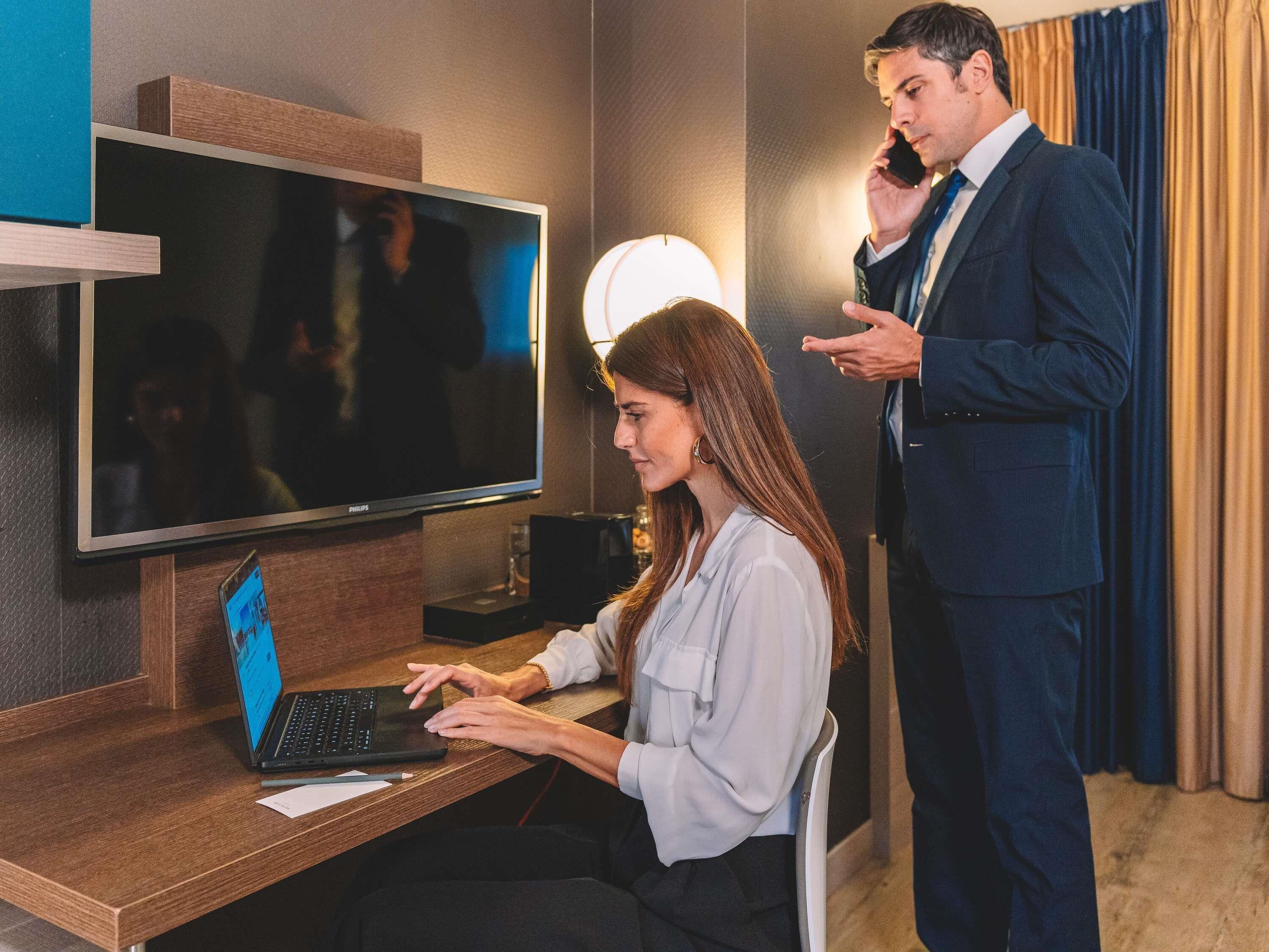 Hotelkamer met een bureau, een laptop, een televisie en een persoon die staat te bellen.