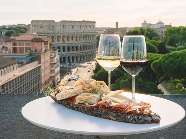 A meal with bread, cheese, and cured meats on a plate, two glasses of wine, and a view of the Colosseum in Rome.