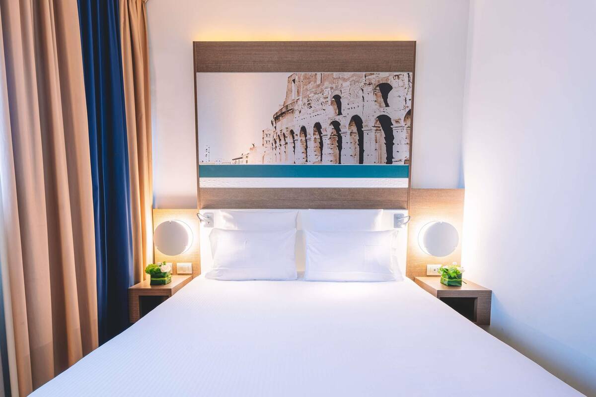Une chambre d’hôtel avec un grand lit, deux tables de chevet dotées de miroirs ronds, une tête de lit ornée d’un tableau représentant un paysage urbain et une vue sur les rideaux.
