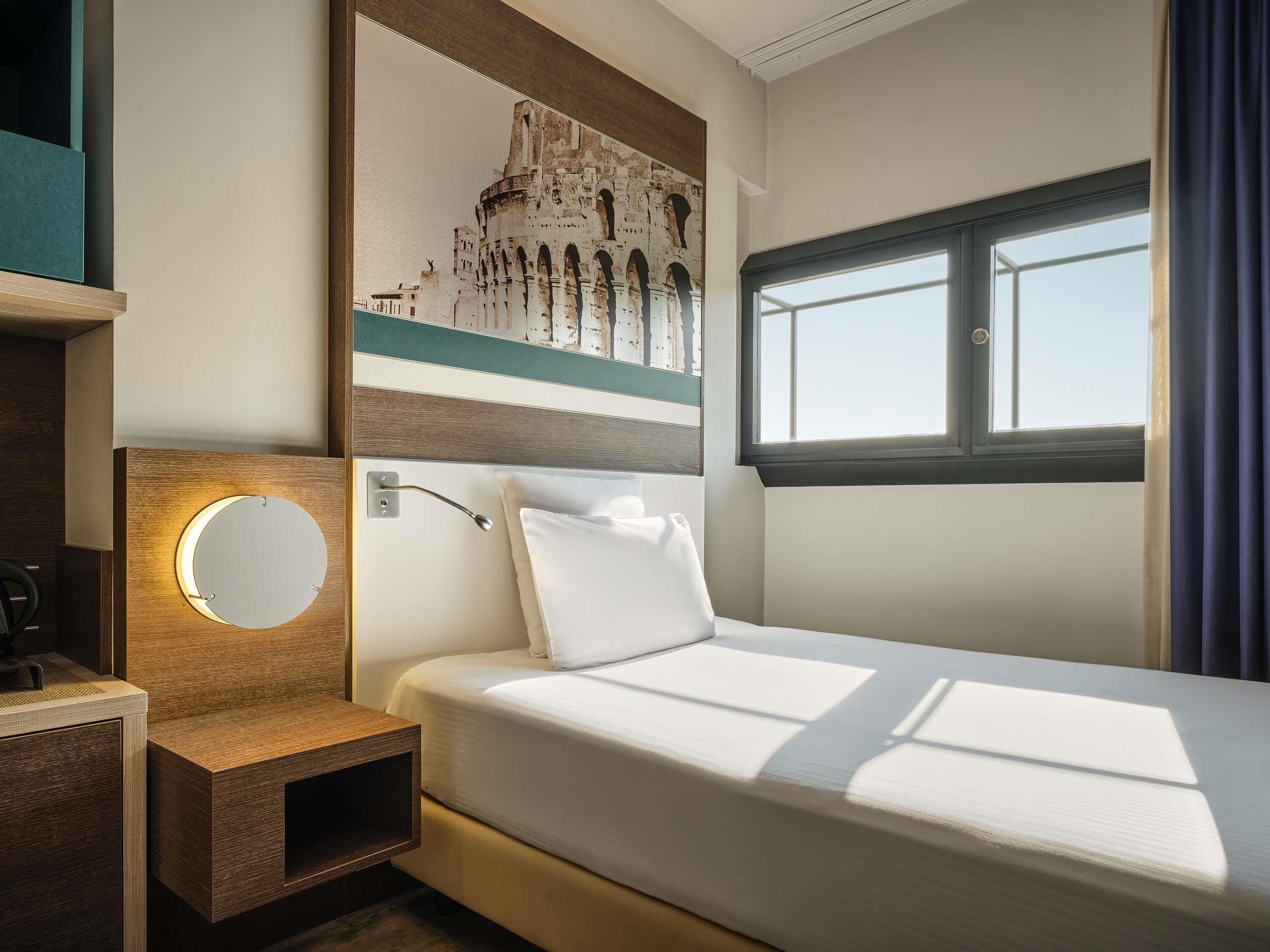 Habitación de hotel con cama, mesita de noche con lámpara, espejo en la pared y un ventanal con cortinas.