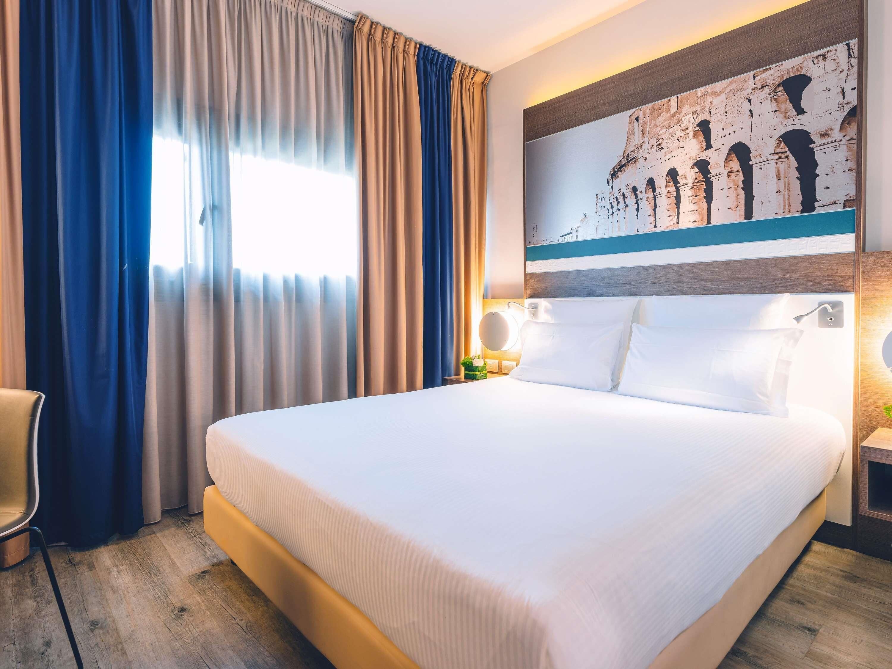 A hotel room with a large bed, a chair, a nightstand with a lamp, and a painting of an architectural structure.
