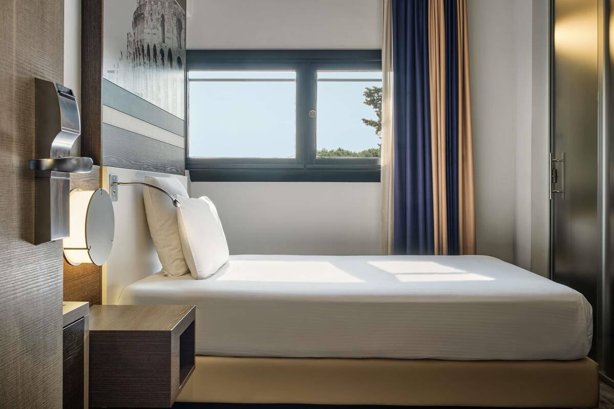 Une chambre d’hôtel comprenant un lit, une table de chevet, une lampe fixée au mur, une fenêtre avec des rideaux et une porte.