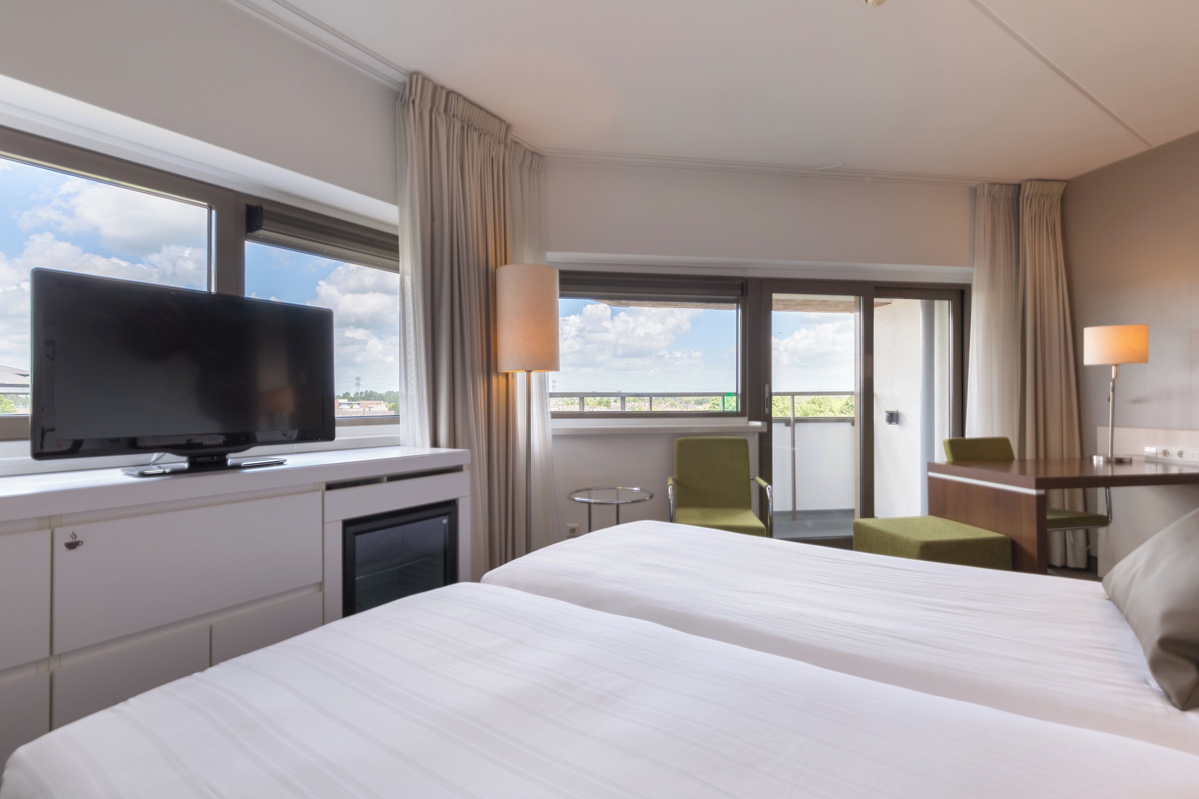 A hotel room with a large bed, a television, a small table, a chair, and a balcony with curtains.