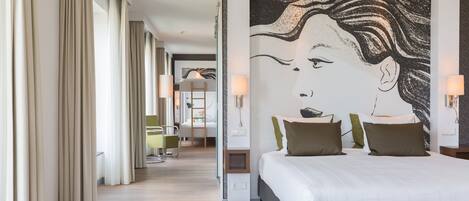 Una habitación de hotel con una cama grande, un mural en la pared y un pasillo con un escritorio y una silla.