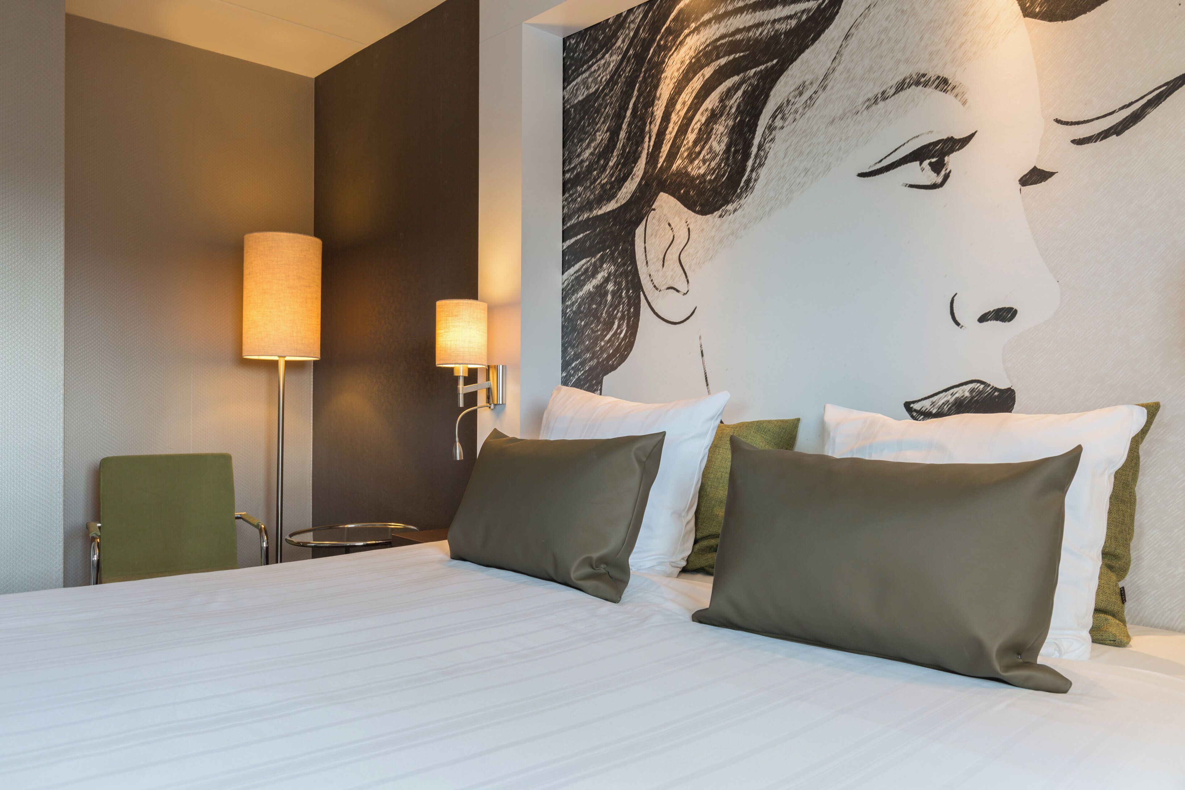 A hotel room with a bed, two wall-mounted lamps, a green chair, and a large abstract painting on the wall.