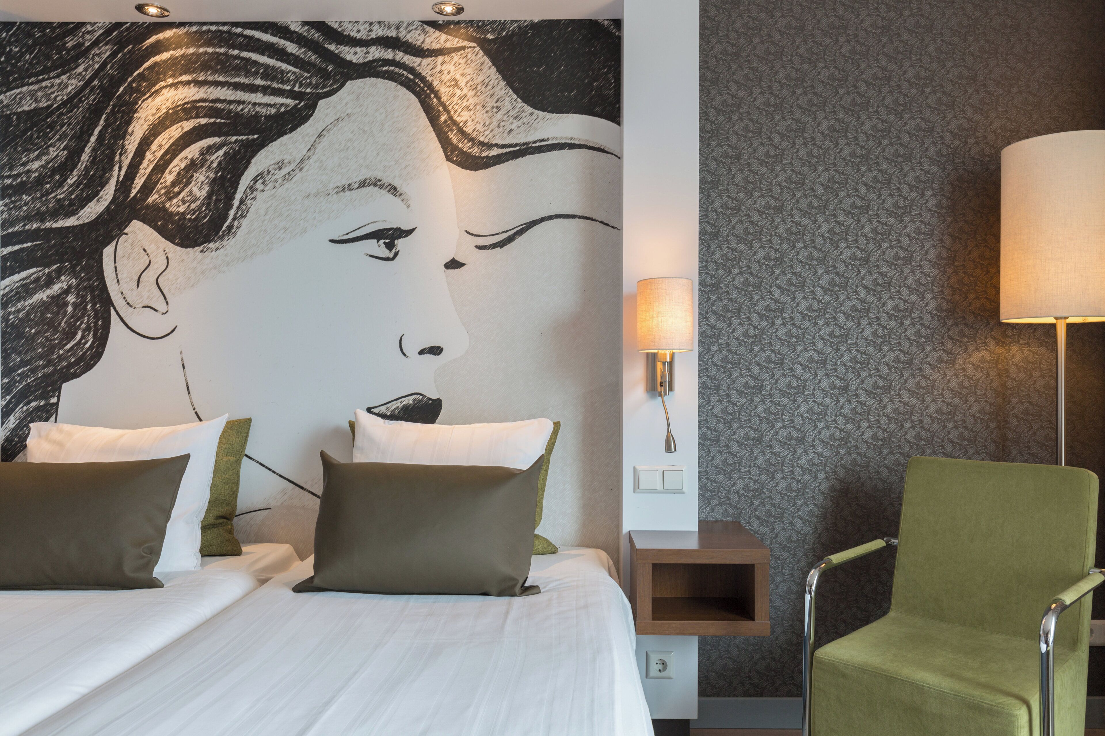 A hotel room with a large bed, a green armchair, a wall-mounted lamp, and a patterned wallpaper.