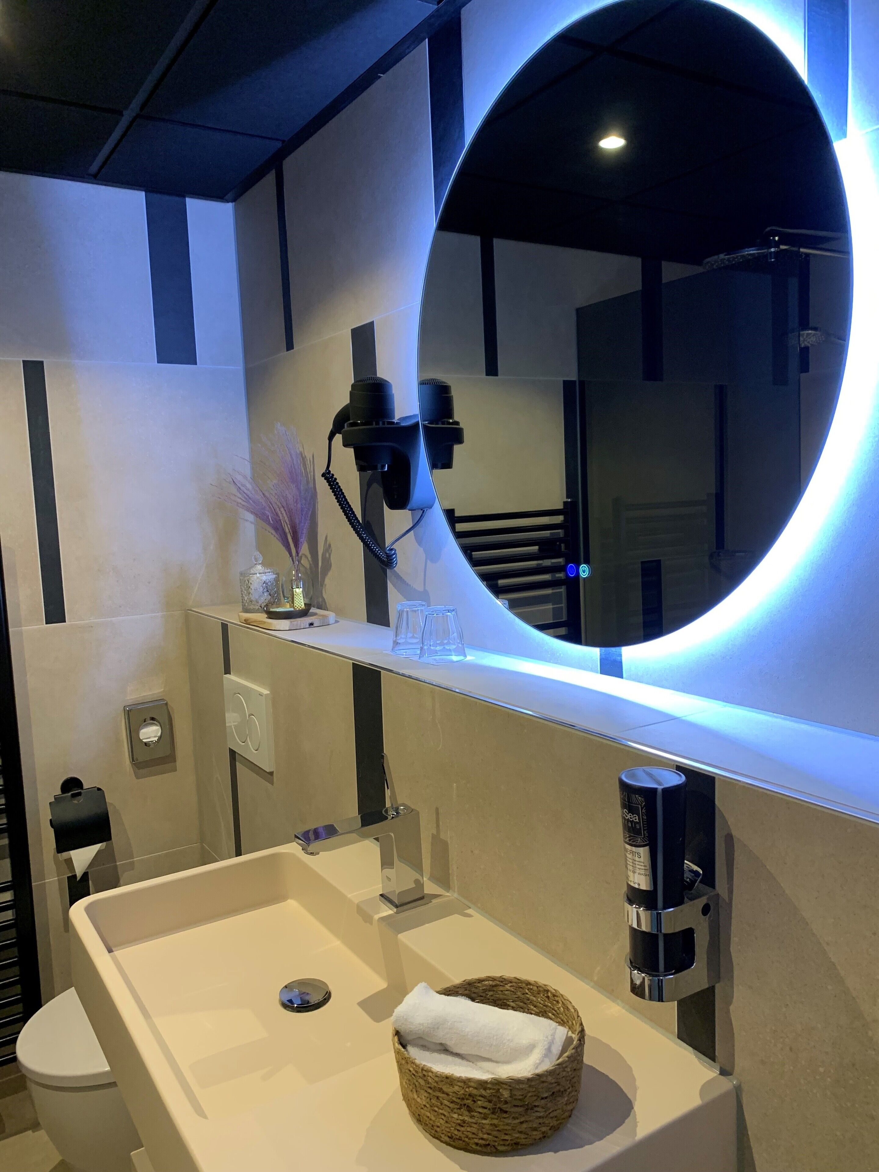A modern bathroom with a large mirror, a white sink, a towel basket, and a shelf with a hairdryer and glasses.