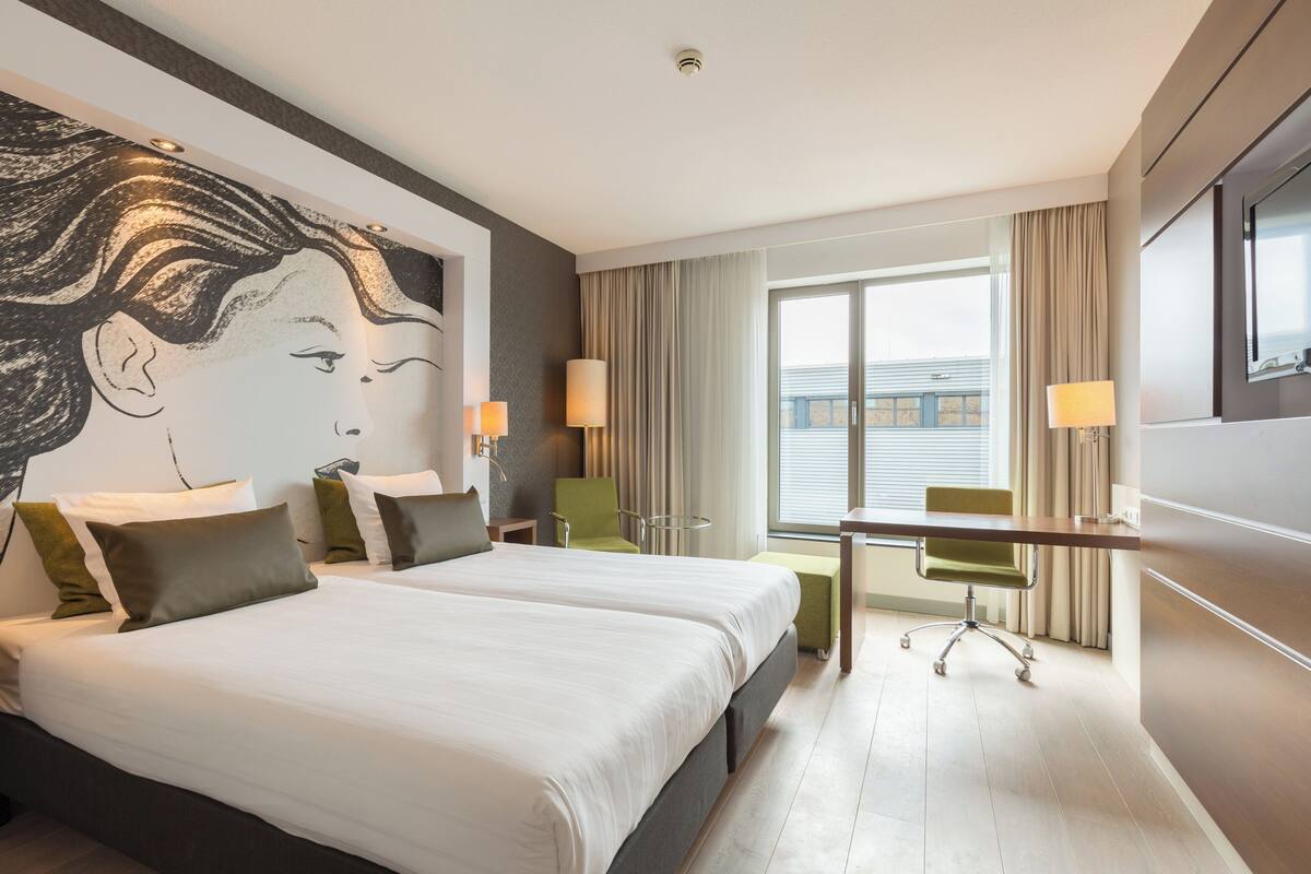 A hotel room with two beds, a desk, and a large window with curtains.