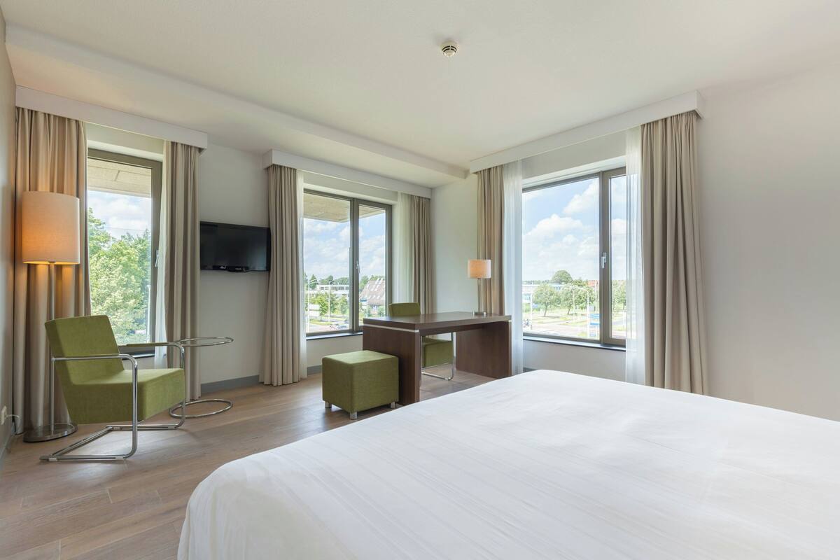 A hotel room with a bed, a desk, two chairs, a television, and large windows with curtains.