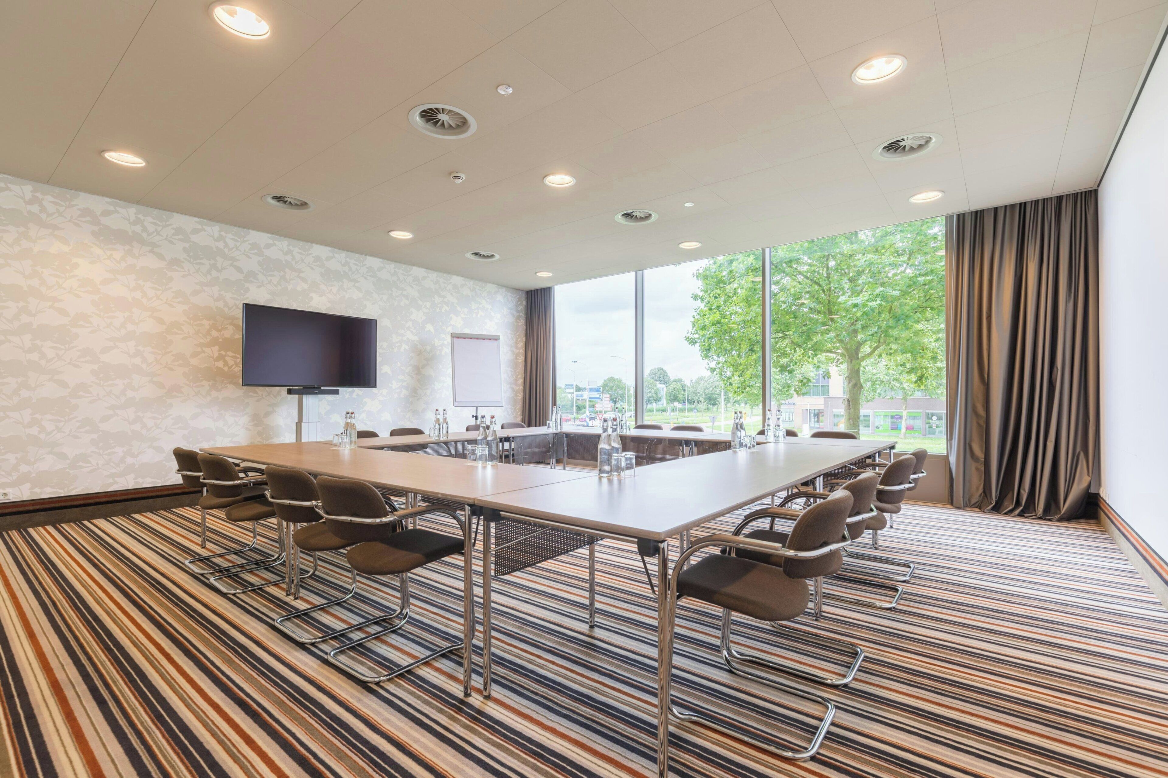A conference room with a long table, multiple chairs, a flat-screen TV, and large windows with curtains.