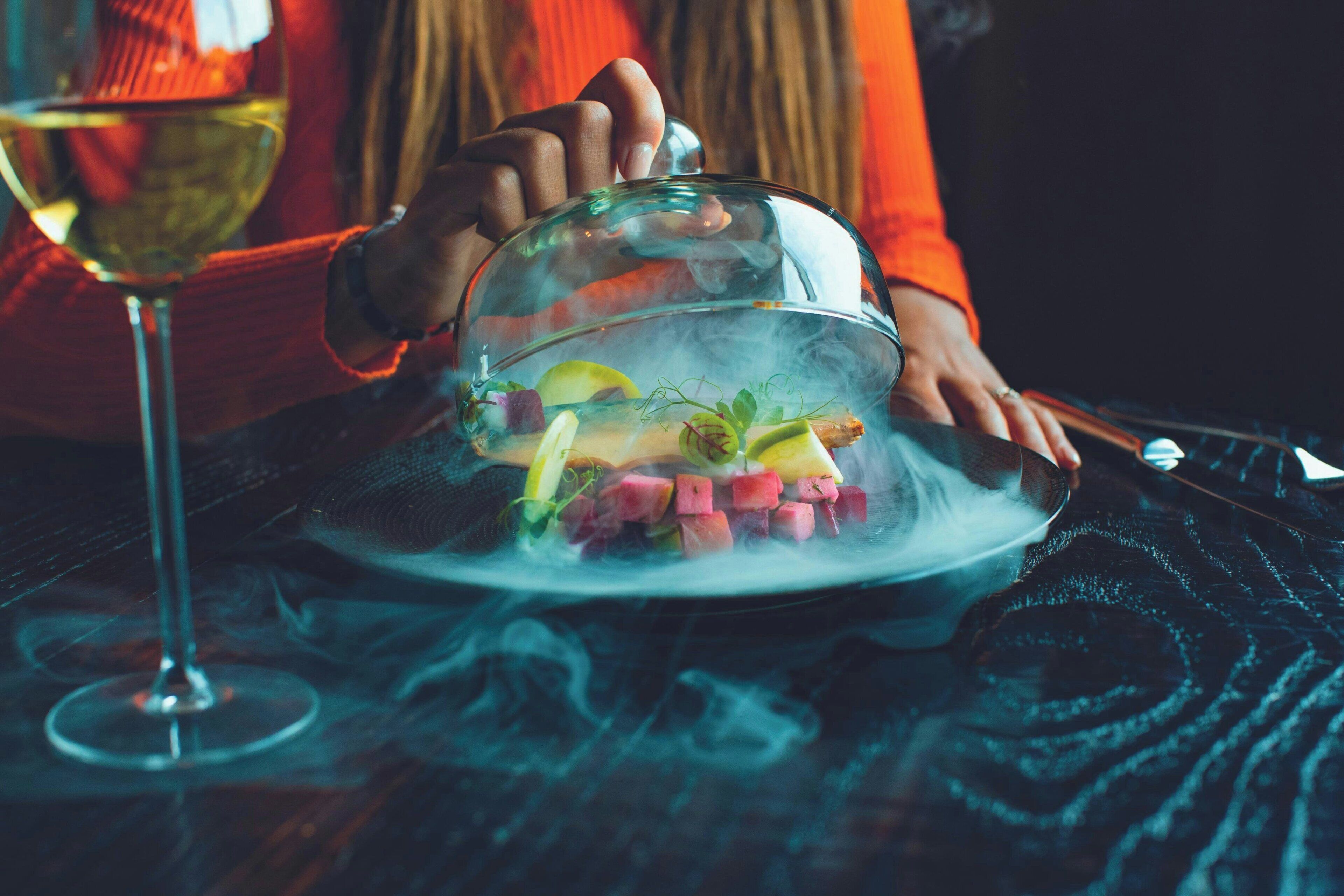 A person holding a glass dome over a dish with food and a glass of wine.