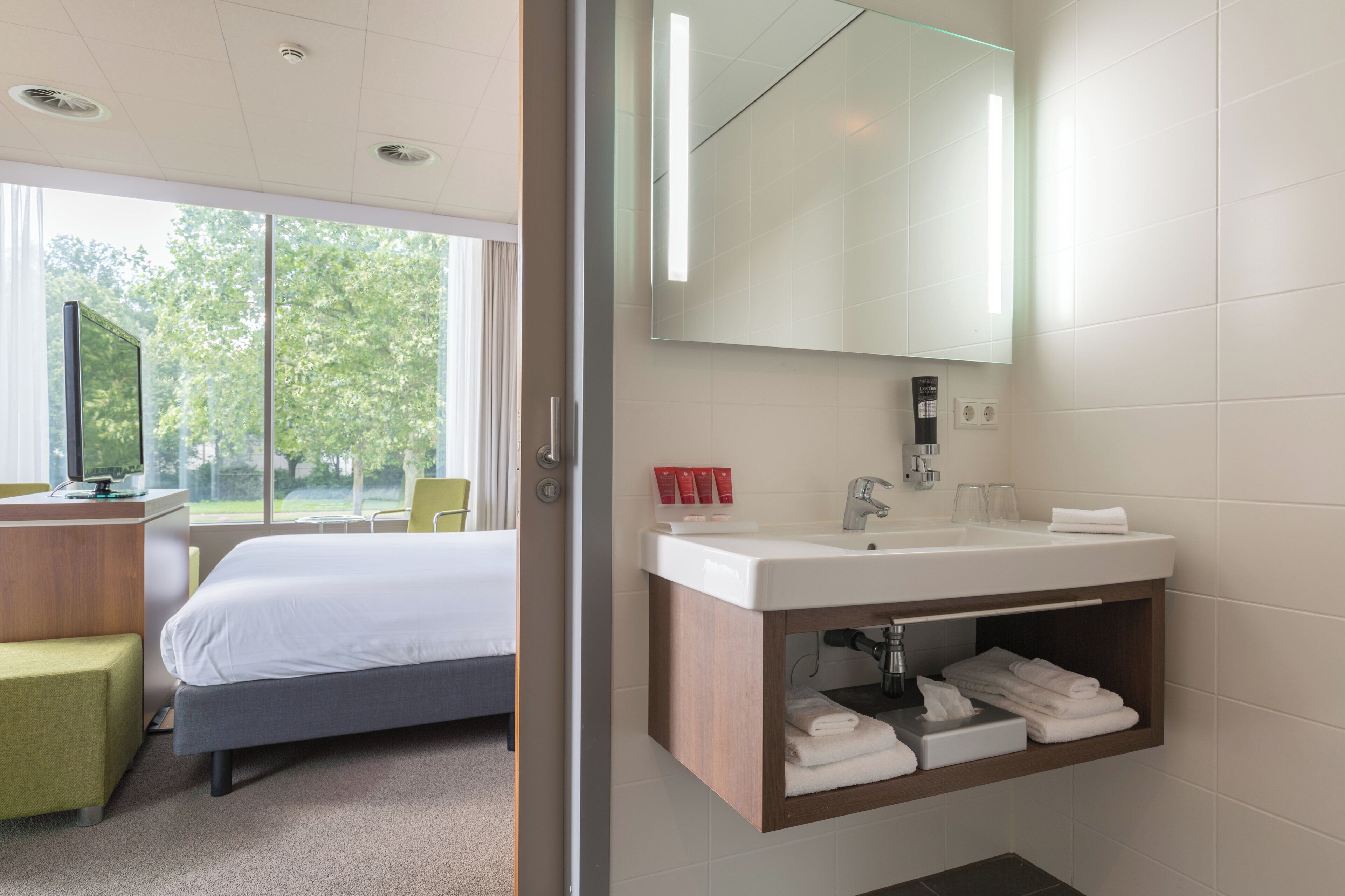 A hotel room with a bed, a desk with a computer, a large window with a view of trees, a bathroom with a mirror, sink, and towel storage.
