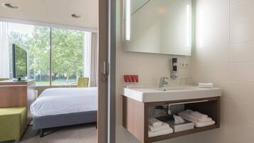 Habitación de hotel con cama, escritorio con computadora, ventana grande con vista a árboles, baño con espejo, lavamanos y espacio para toallas.