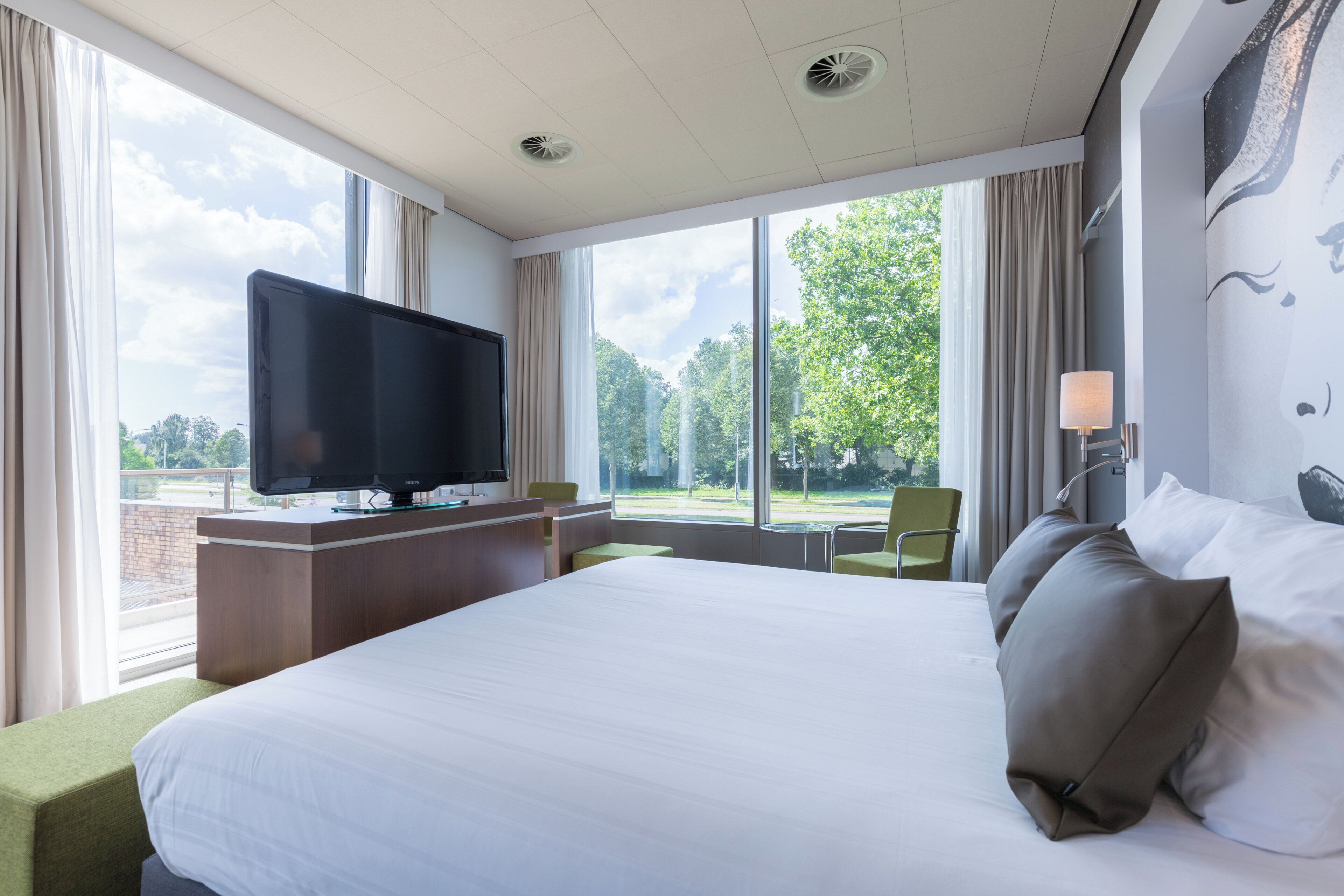 Habitación de hotel con una cama grande, un televisor de pantalla plana, un escritorio y vistas a árboles a través de amplias ventanas.