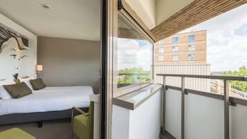 Hotellihuone, jossa on sänky, pieni pöytä, tuoli ja parveke, jolta on näkymä rakennukseen ja puihin.