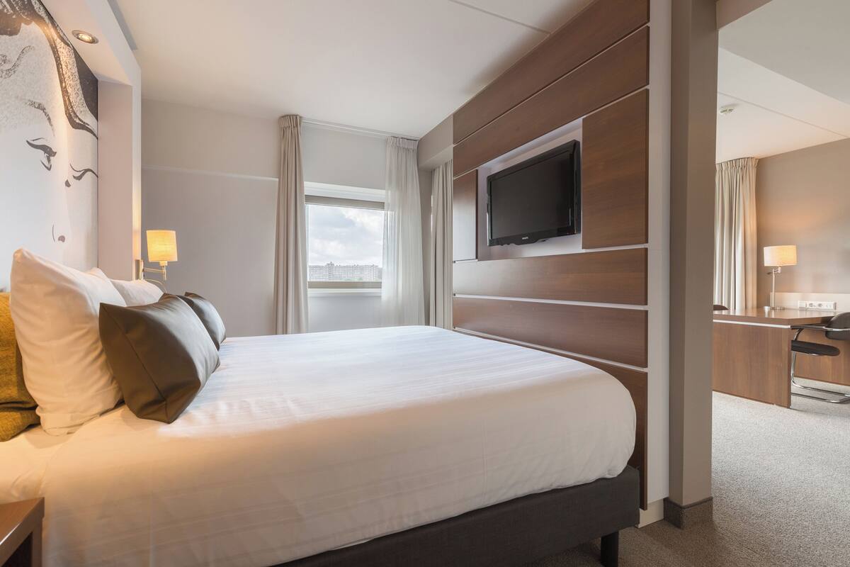 A hotel room with a large bed, a flat-screen TV mounted on the wall, a desk with a chair, and a window with curtains.