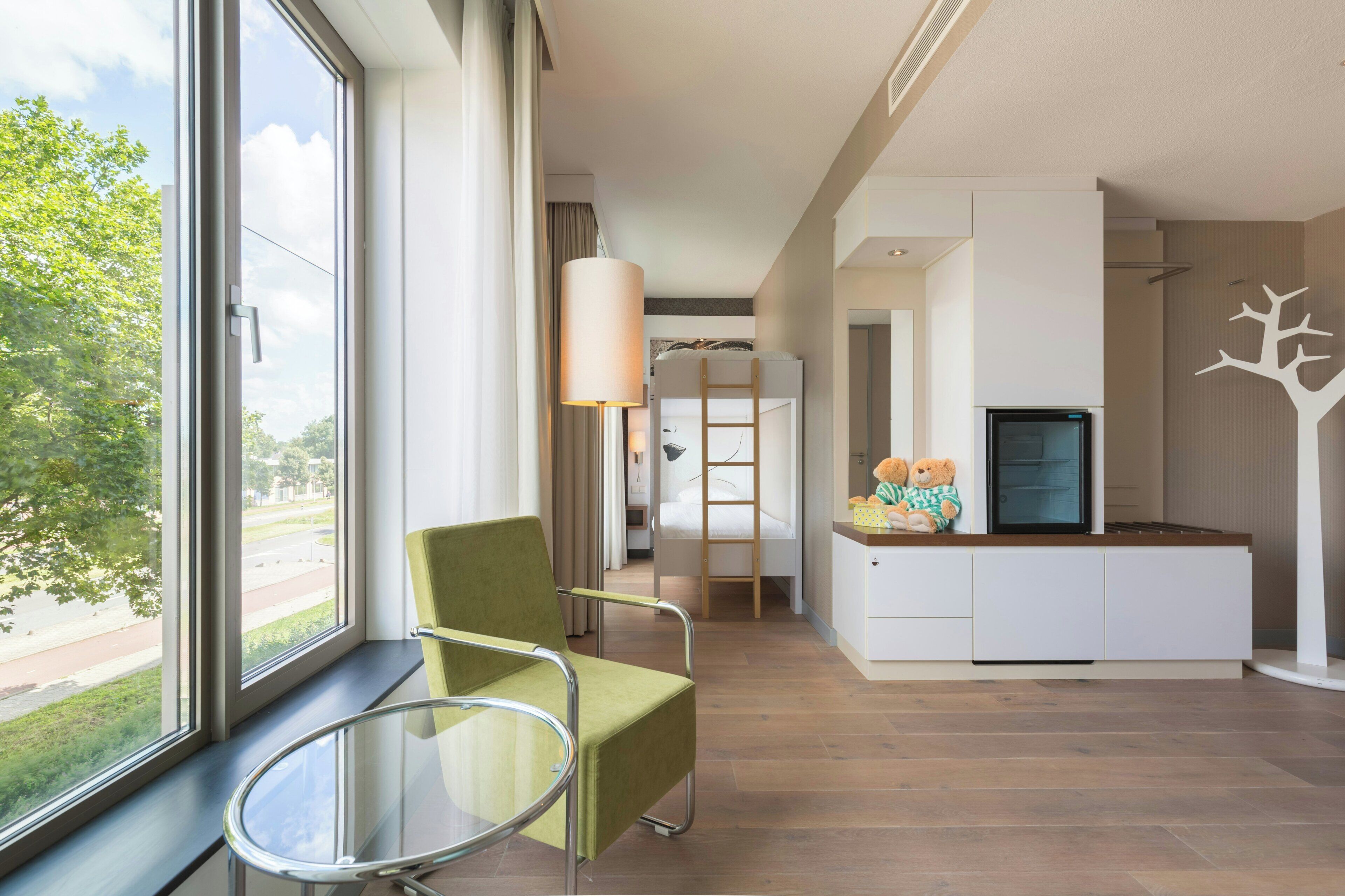 Una habitación de hotel moderna con un ventanal grande, una silla verde, una mesita pequeña, un armario blanco y un televisor.