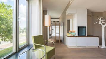 A modern hotel room with a large window, a green chair, a small table, a white cabinet, and a TV.
