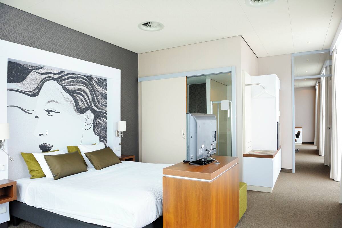 A modern hotel room with a large bed, a desk with a computer, a mirror, and a view of the hallway.