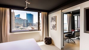 Een moderne hotelkamer met een groot raam met uitzicht over de stad, een opgemaakt bed, een klein voetenbankje en een bureau met stoel.