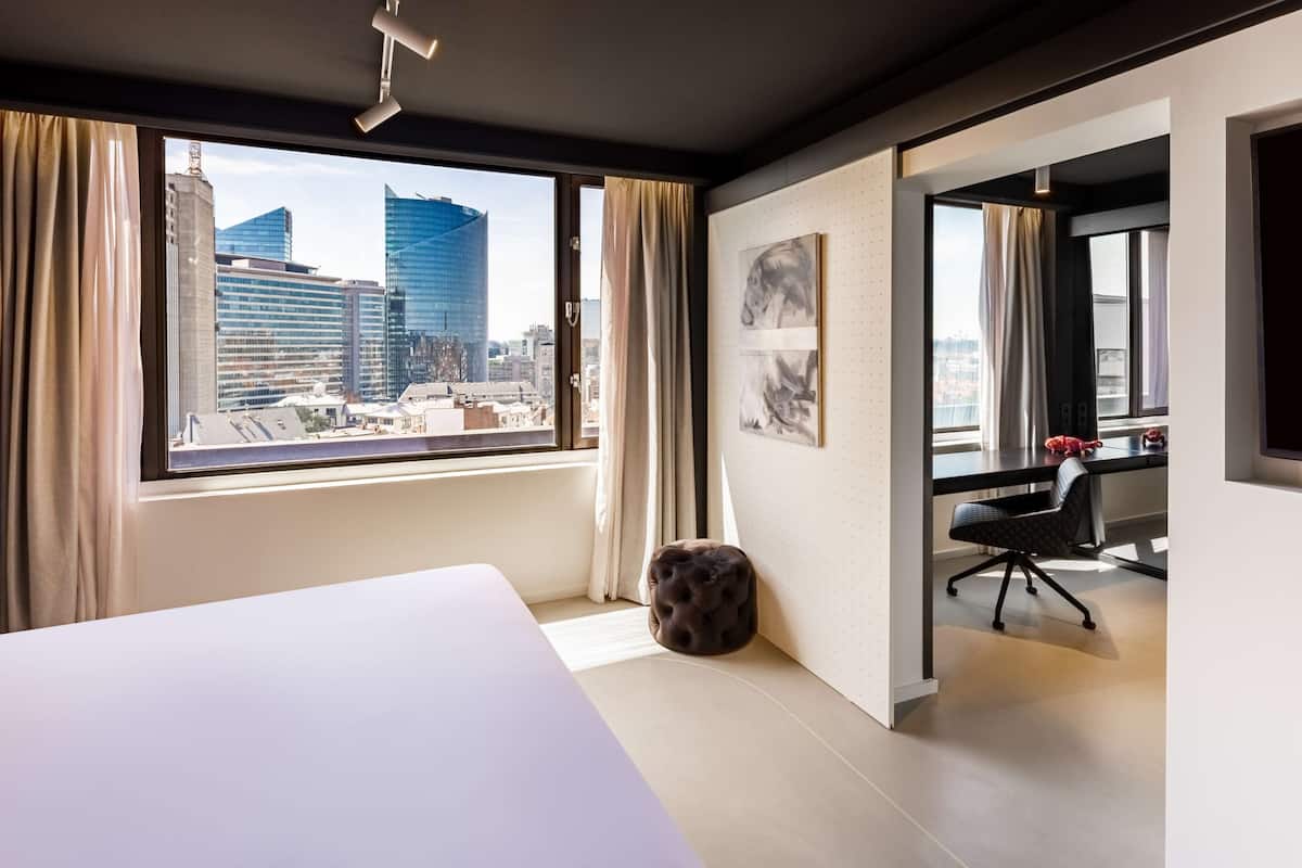 A modern hotel room with a large window offering a city view, a neatly made bed, a small ottoman, and a desk with a chair.