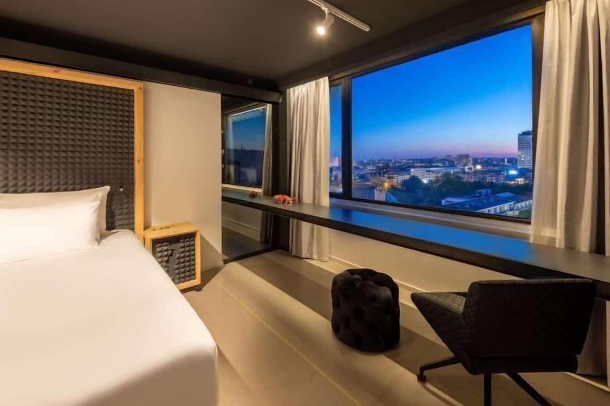 A hotel room with a bed, a desk with a chair, a large window with a city view, and a soundproof wall.