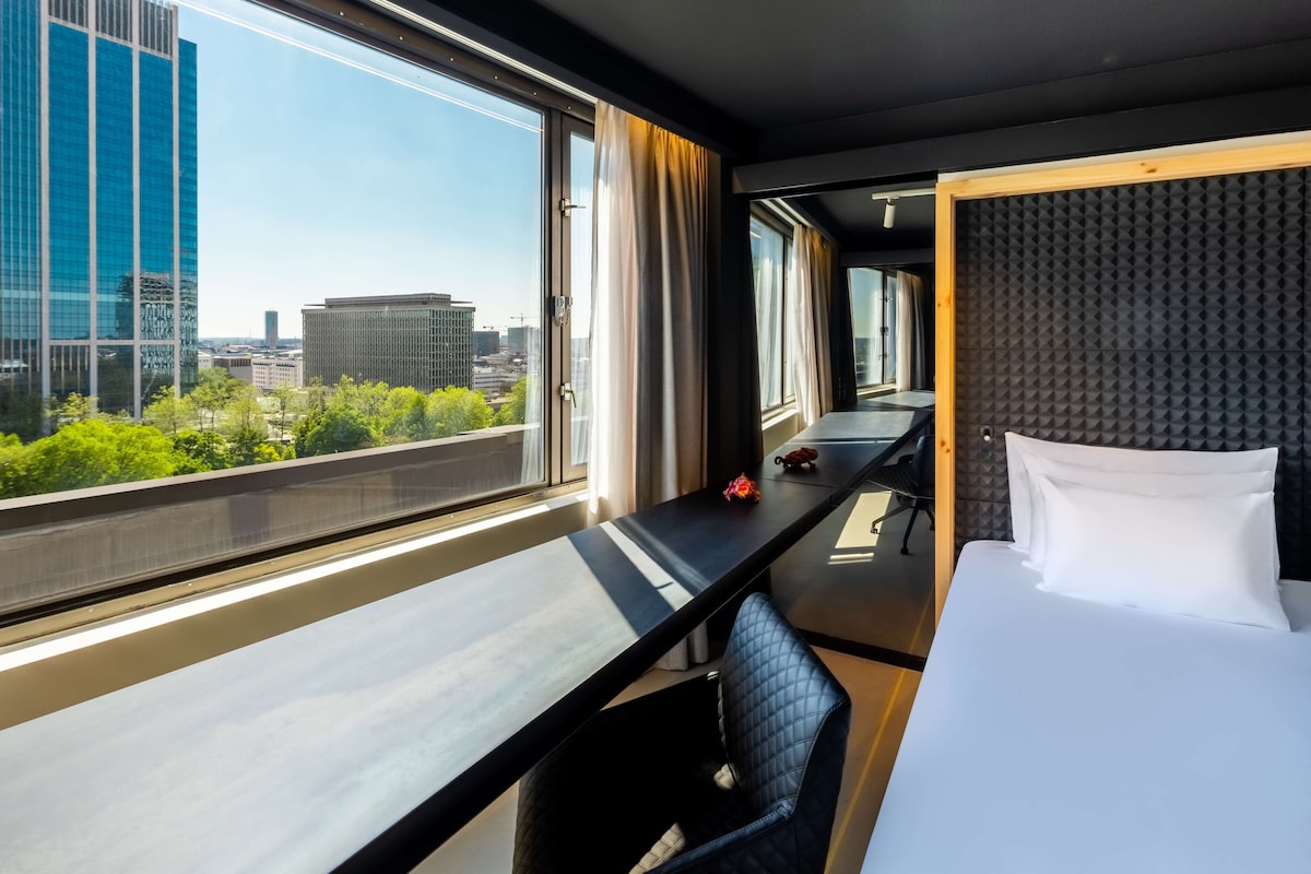 A hotel room with a large window offering a city view, a bed with white linens, a desk with a chair, and a soundproof wall.