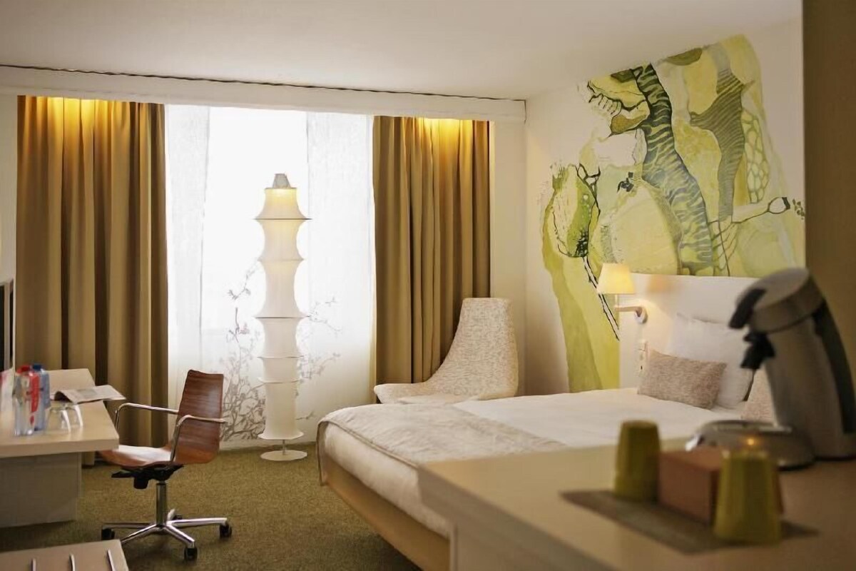 A hotel room with a bed, a desk, a chair, a lamp, and a painting on the wall.