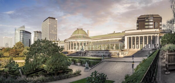 A historic building with a dome and classical architecture, surrounded by a well-maintained garden and modern skyscrapers in the background.