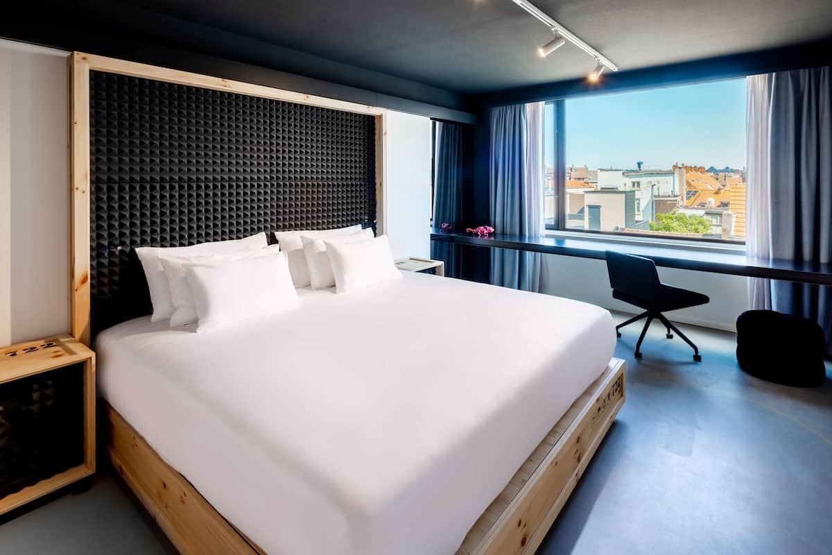 A modern hotel room with a large bed, a wooden headboard, a black office chair, and a view of the city through large windows.