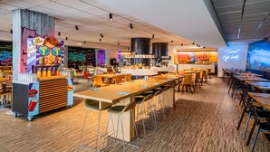 Een moderne cafetaria met lange houten tafels en stoelen, een kleurrijke muurschildering en een neonbord.