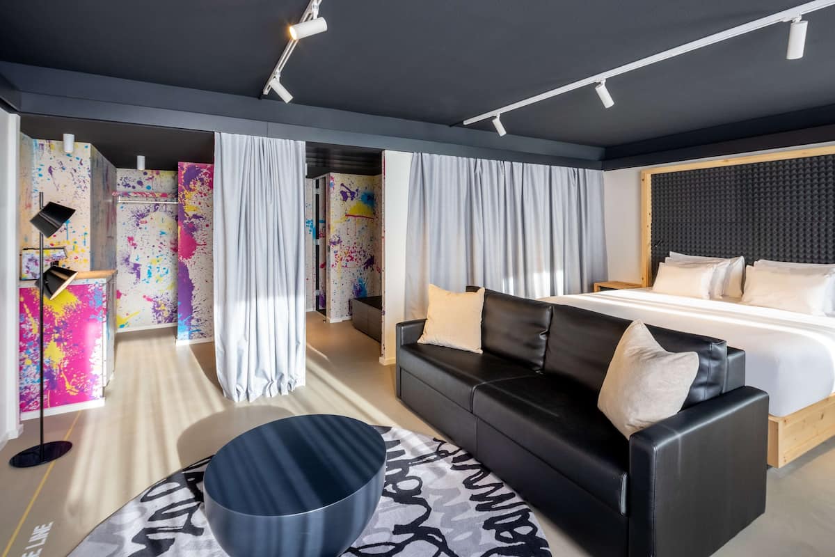 A modern hotel room with a black leather sofa, a bed with white bedding, and colorful abstract wall art.
