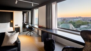 Een moderne hotelkamer met een groot raam dat uitzicht geeft op de stad, een bureau met stoel, een bank en een klein voetenbankje.