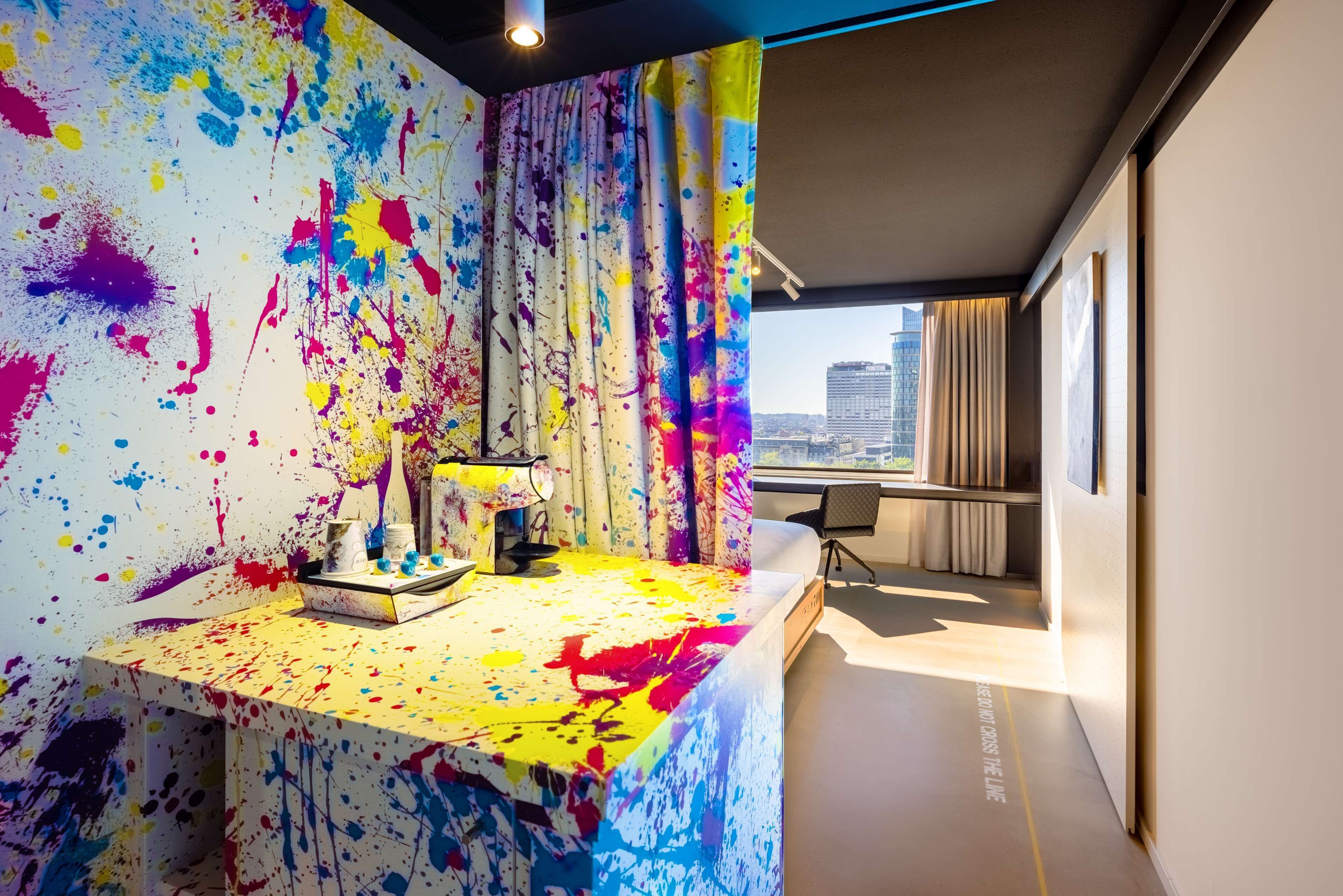 A modern room with a vibrant abstract painting on the wall, a desk with a lamp, and a view of a cityscape through large windows.