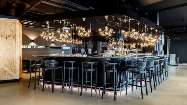 Een moderne bar met een lange toonbank, meerdere krukken en hangende bolvormige lampen.