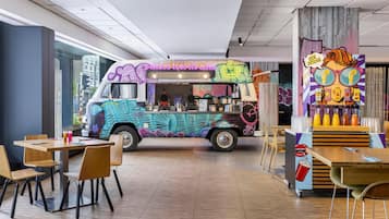 Un camión de comida con grafitis de colores, un espacio de descanso con sillas y mesas de madera, y un mural en la pared.