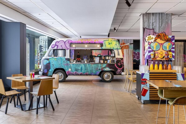 Un camión de comida con grafitis de colores, un espacio de descanso con sillas y mesas de madera, y un mural en la pared.