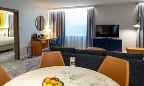 Habitación de hotel con cama, escritorio, televisor, sofá y mesa de comedor con fruta.