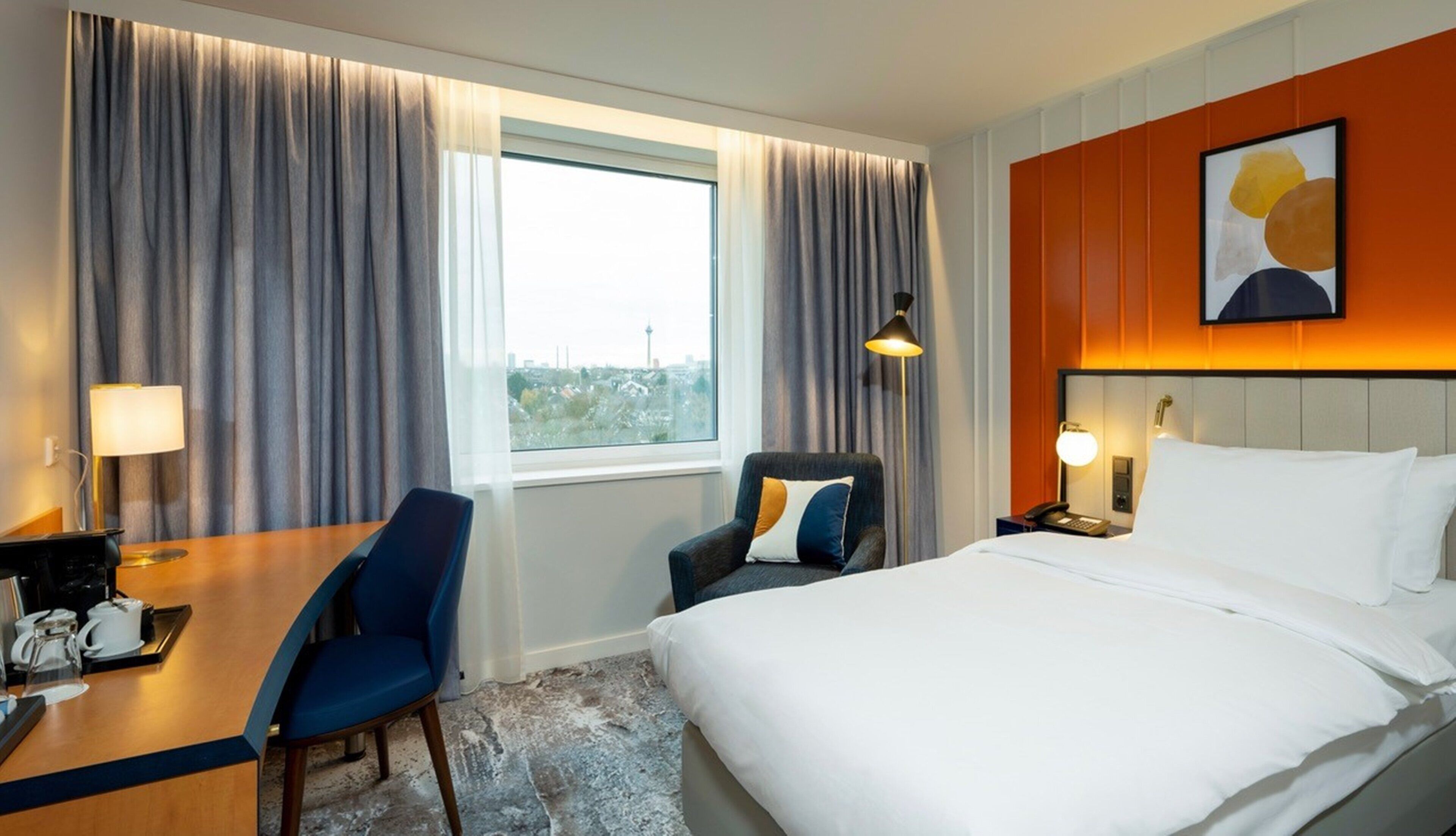 A hotel room with a bed, a desk with a chair, a telephone, and a window with curtains.