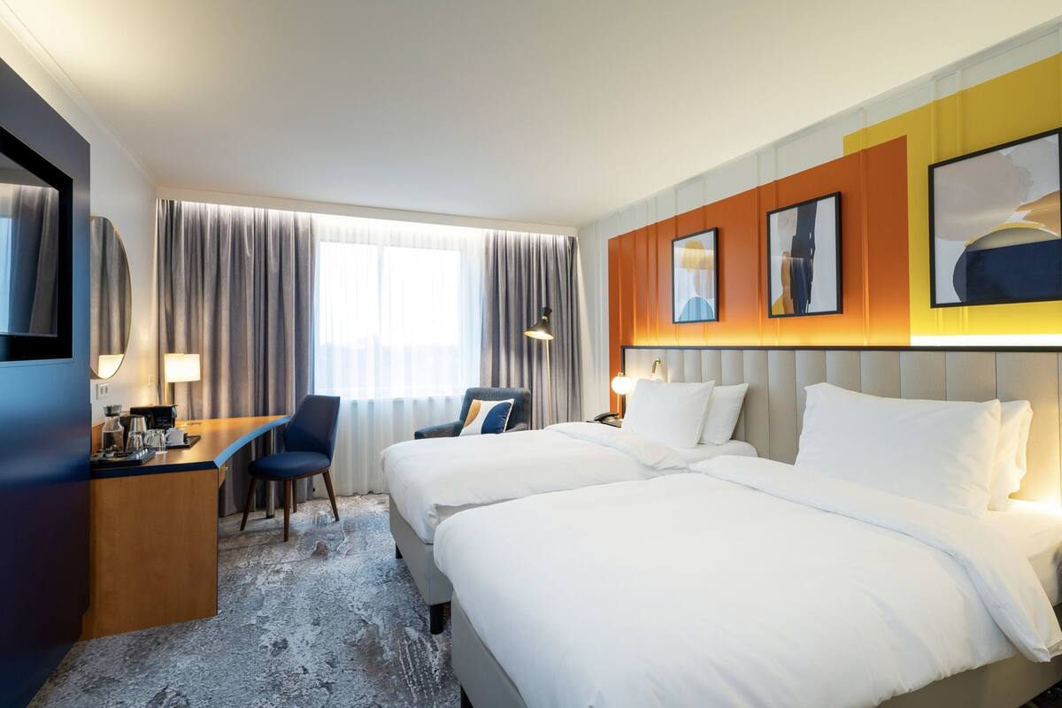 A hotel room with two beds, a desk, a chair, a TV, and a window with curtains.