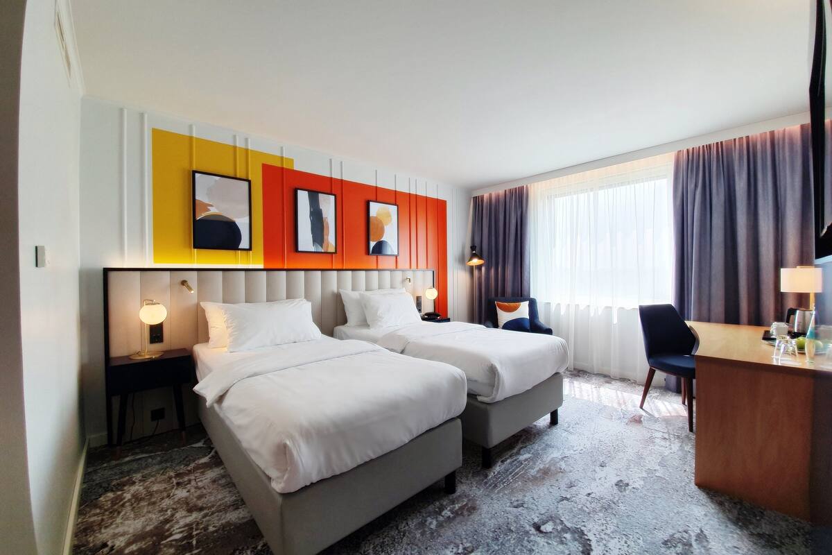 A hotel room with two beds, a desk, and a chair. There are wall-mounted lamps and a large window with curtains.