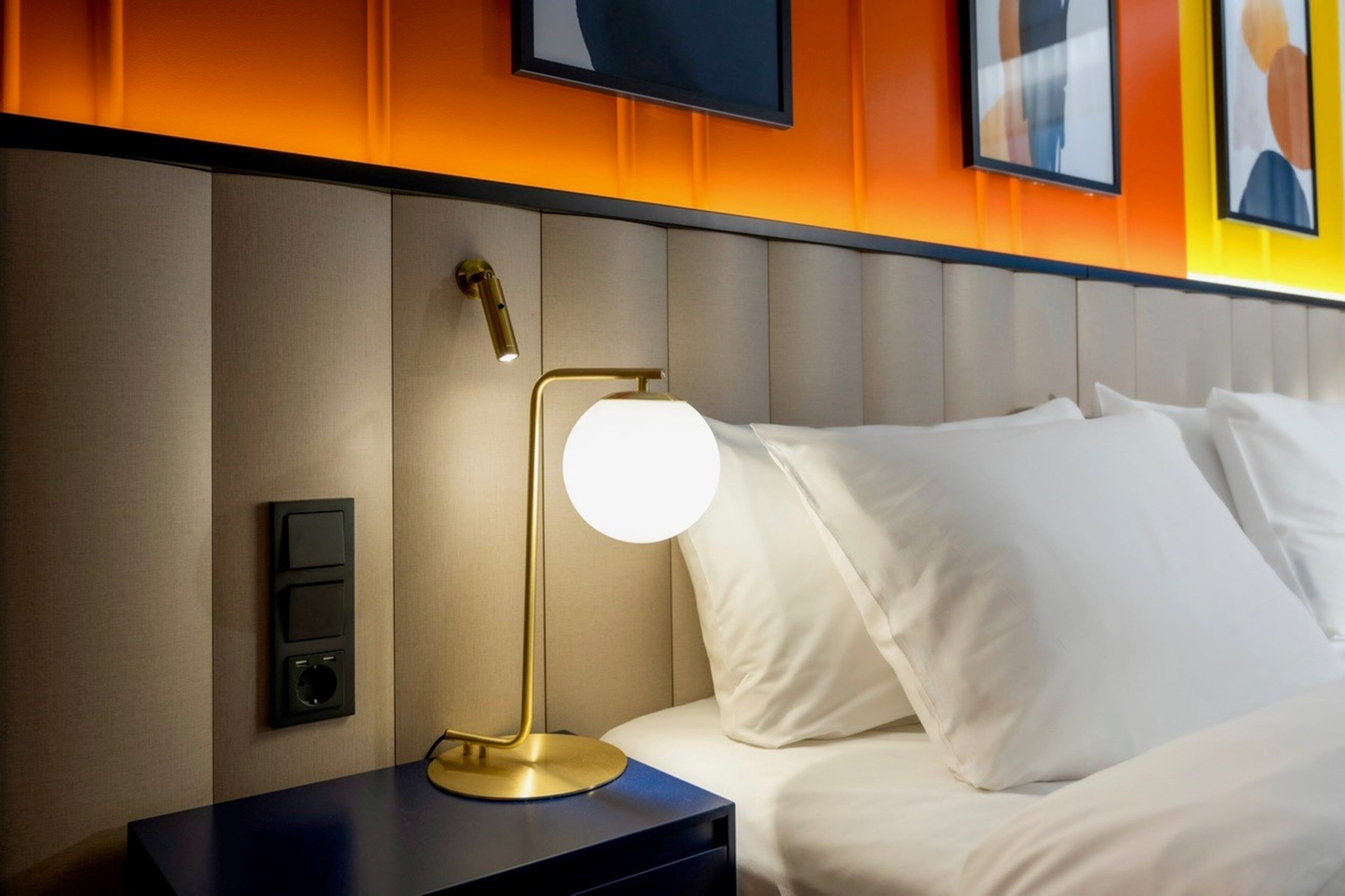 A neatly made bed with white linens, a bedside table with a lamp, and a wall-mounted control panel.