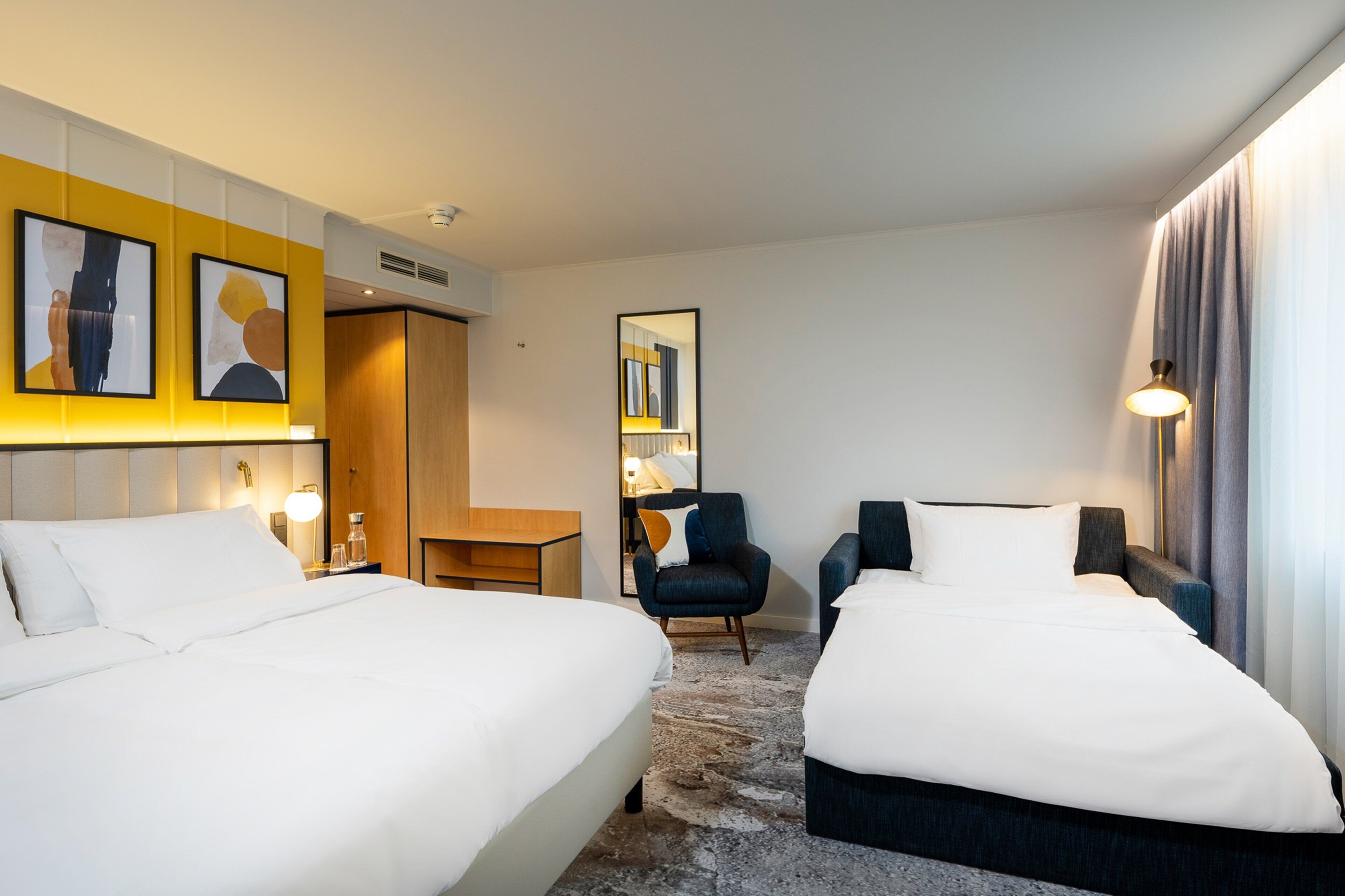 Hotellihuone, jossa on kaksi sänkyä, työpöytä, tuoli, peili ja lamppu.