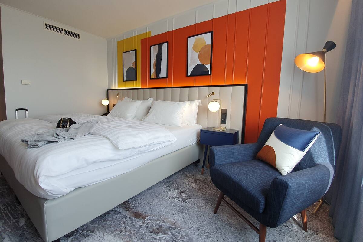 A hotel room with a bed, a chair, a nightstand, and a wall with three framed pictures.