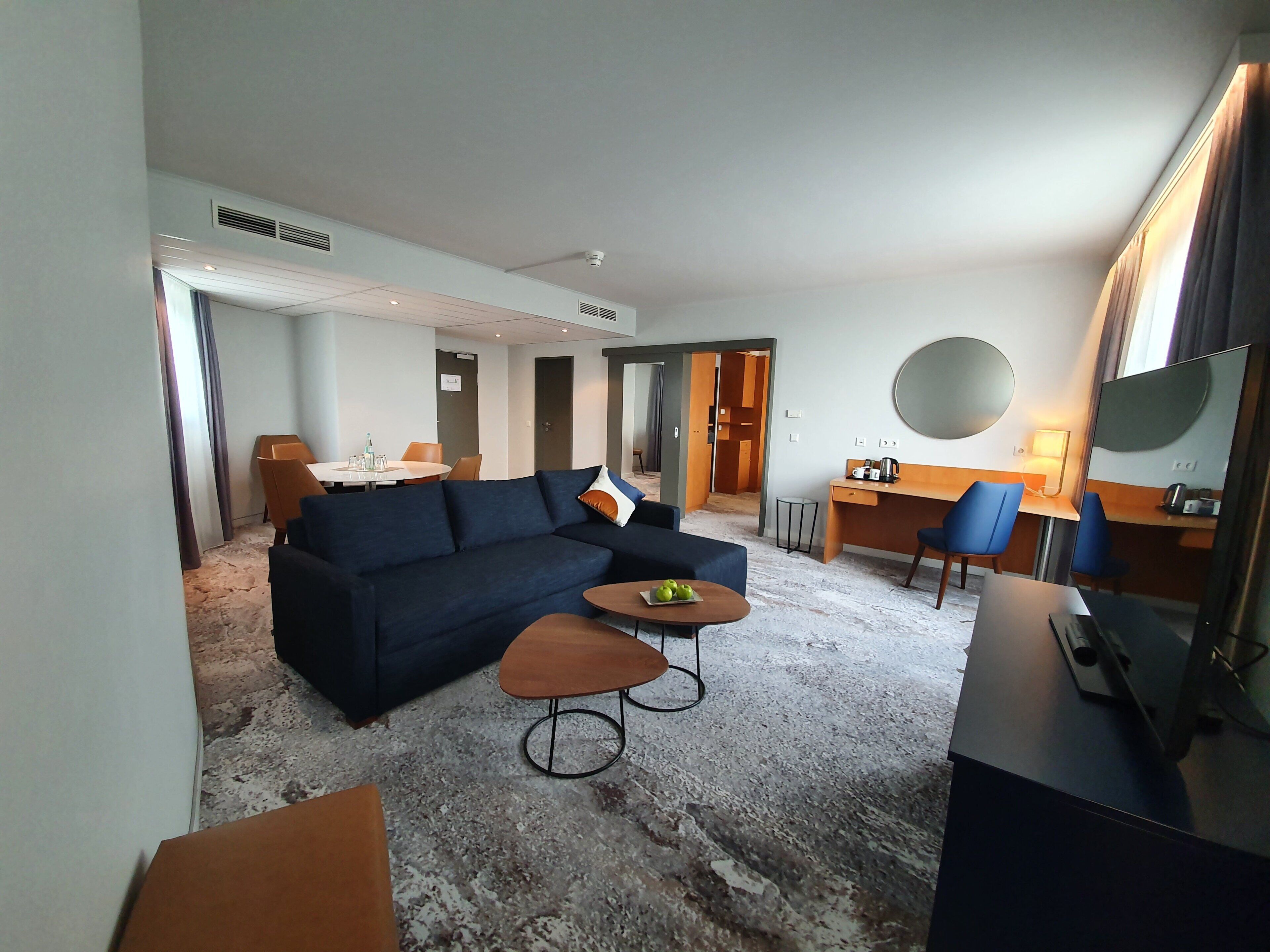 A hotel room with a dark blue sofa, a small round table with apples, a desk with a chair, a TV, and a large mirror.