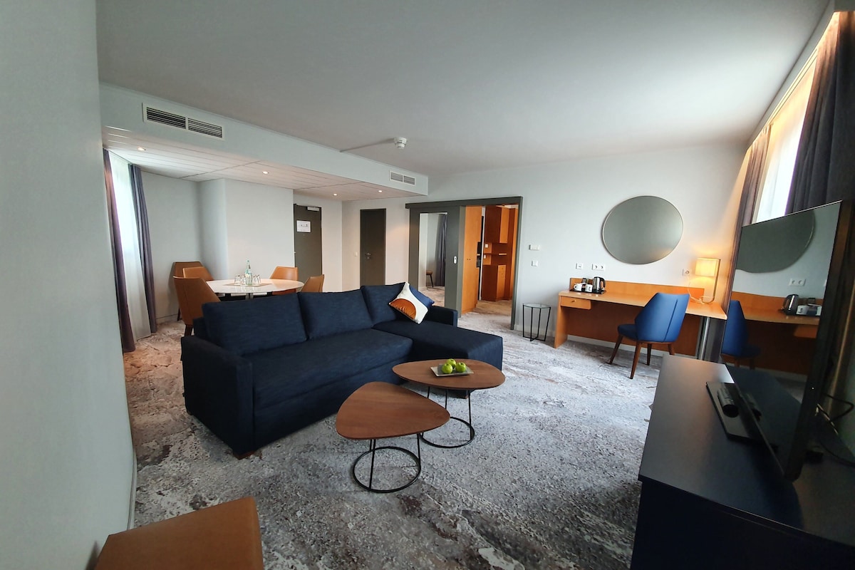 A hotel room with a dark blue sofa, a small round table with apples, a desk with a chair, a TV, and a large mirror.